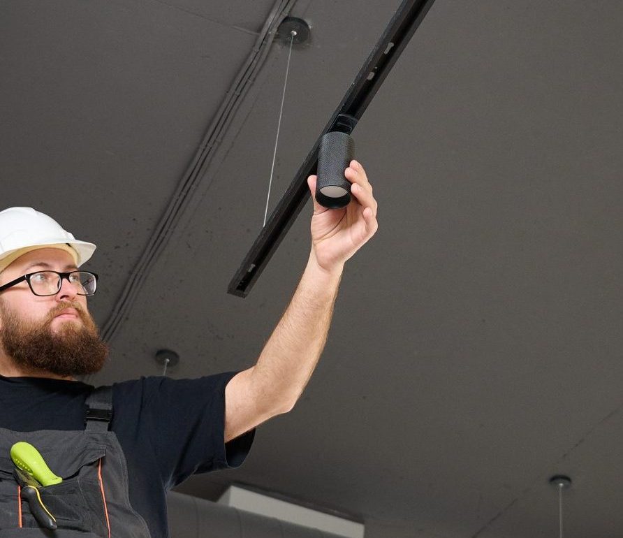 Man install a spot track LED light in the ceiling at the new office. Electrician staying on ladder. Industrial black loft design. Bearded man installing track light at the new office