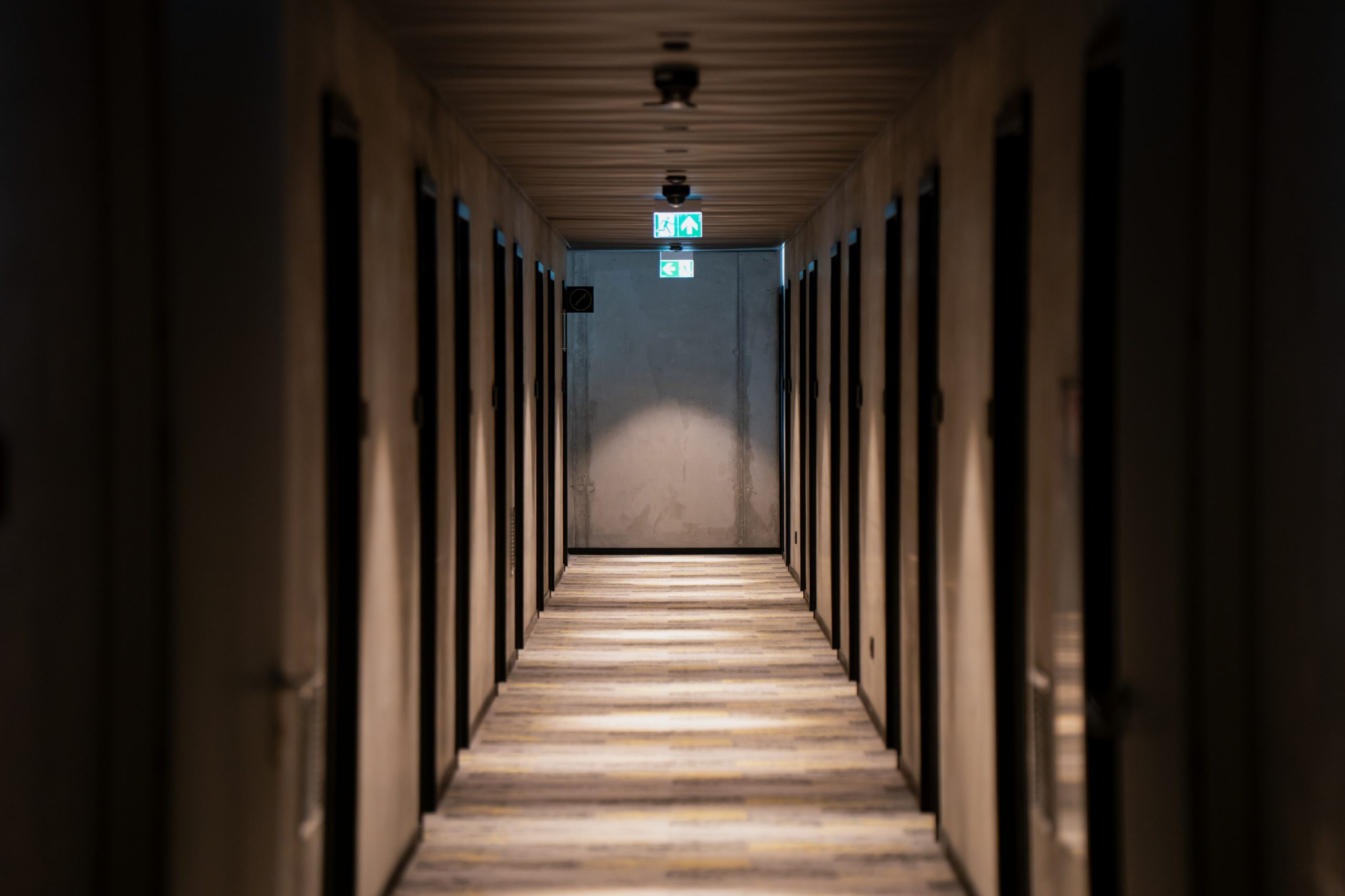 Interior hallway with closed wooden doors and emergency exit sign in industrial or commercial building with minimal lighting and symmetrical architectural design