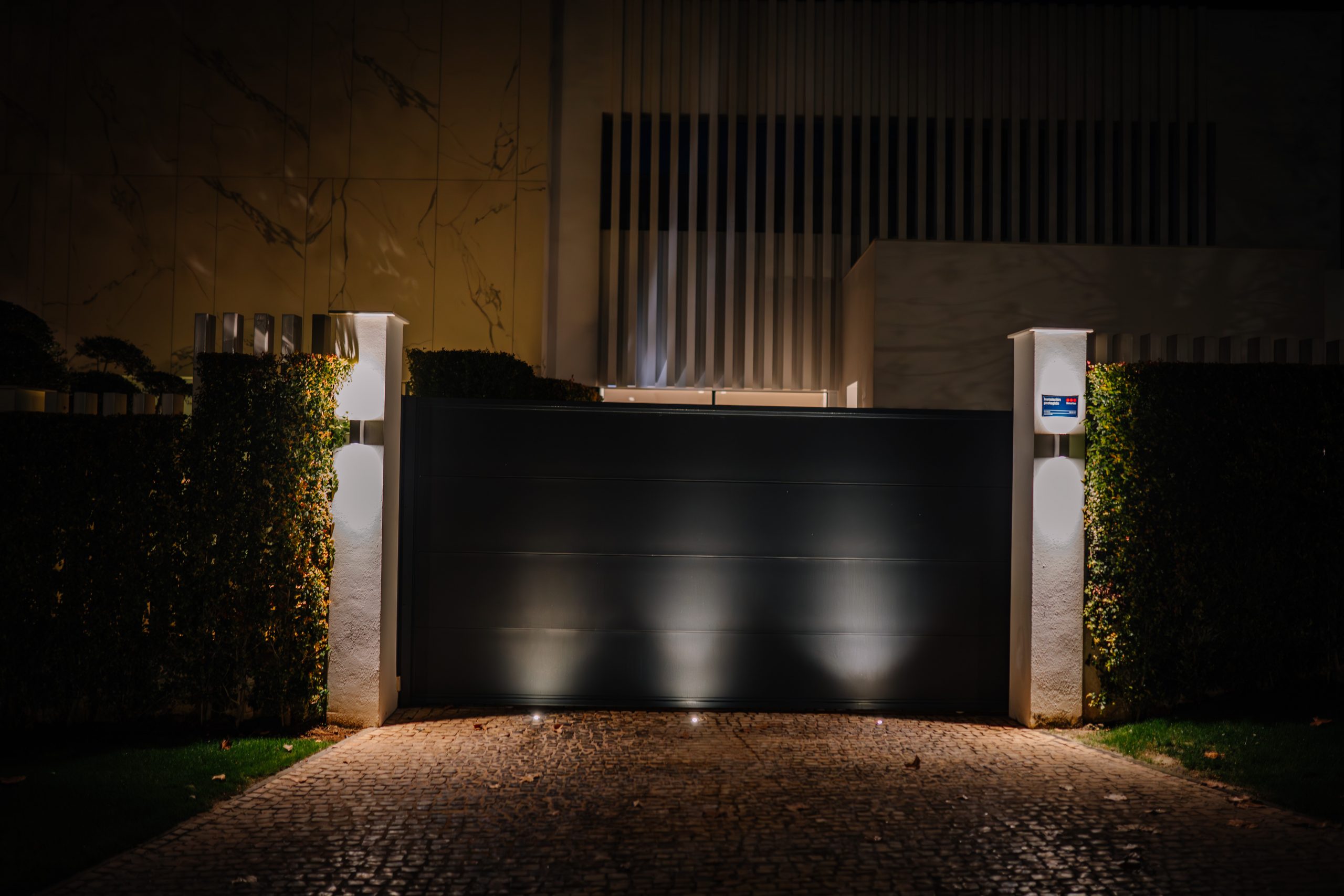 A night view of a luxury entrance with a black sliding gate, white walls, and soft lighting.